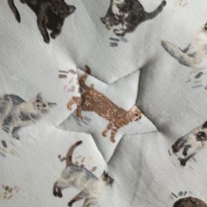 May include: Close-up of a light gray fabric with a repeating pattern of various cat illustrations in brown, black, and gray. The fabric is quilted with a star-shaped pattern.