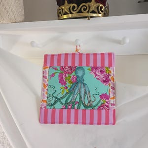 May include: A square fabric potholder featuring a central panel with a teal octopus surrounded by pink and white floral patterns. The top and bottom borders consist of vertical pink and white stripes, while the side borders feature orange leaves.