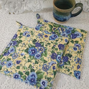 May include: Two square quilted pot holders featuring a yellow background with a repeating pattern of blue and purple pansy flowers and green leaves. The fabric has a stitched grid texture, and each pot holder includes a small fabric loop for hanging.