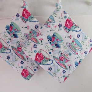 May include: Two square pot holders with a white background and a floral pattern featuring teacups, roses, and small flowers in shades of pink, blue, and green. Each pot holder has a loop for hanging.