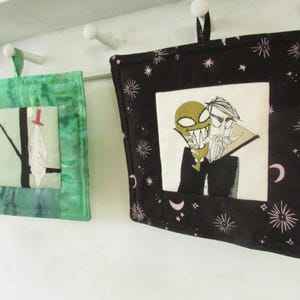 May include: Two square quilted pot holders. One is green with a tree and figure design. The other is black with a cartoon vampire design and a star and moon pattern. Both have hanging loops.