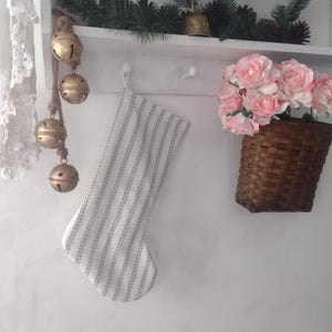 May include: A white stocking with a green and white striped pattern hangs on a white shelf with a garland of pine branches and gold bells. A basket of pink roses sits on the shelf below.