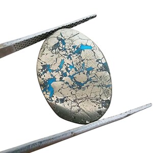Può includere: Una gemma ovale tenuta da una pinzetta. La pietra presenta una superficie argentata metallica screpolata con vivaci venature blu turchese. La gemma è posta su uno sfondo bianco pulito, evidenziando il suo colore e la sua consistenza unici.