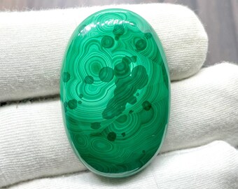 May include: A green malachite cabochon with a swirling pattern of concentric circles and dark green spots. The cabochon is oval shaped and has a smooth, polished surface.