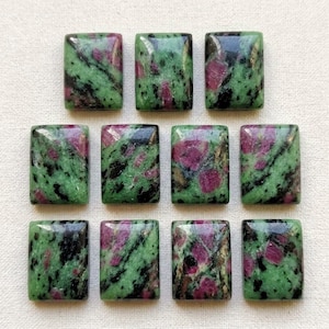 Puede incluir: Once cabujones rectangulares de zoisita con inclusiones de rubí, dispuestos en tres filas. Cada piedra presenta una superficie pulida con un patrón moteado de zoisita verde vibrante, parches de rubí rojo magenta intenso y hornblenda negra.