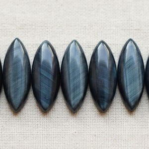 May include: Seven marquise-shaped blue tiger's eye gemstones arranged in a horizontal row. Each stone features a smooth, polished surface with a distinct chatoyant sheen, displaying dark blue and grey linear patterns across the elongated oval shape.