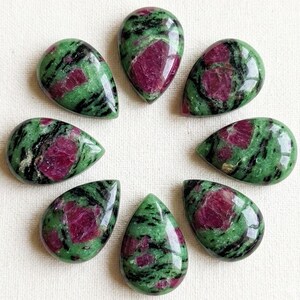 Puede incluir: Siete cabujones pulidos en forma de lágrima de zoisita con rubí. Cada piedra presenta una base verde vibrante con inclusiones de hornblenda negra y manchas de rubí rojo magenta opaco, dispuestas en un patrón circular.