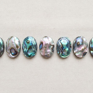 Può includere: Sette cabochon in conchiglia di abalone di forma ovale disposti in una fila orizzontale. Ogni pezzo presenta una superficie iridescente unica con motivi vorticosi nei toni del blu, verde, viola e rosa, con una finitura liscia e lucida.