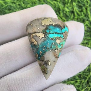 Op de afbeelding: Een druppelvormige edelsteen met een mix van kleuren. De steen heeft een basis van lichtgrijs en wit, met aderen van turquoise en gouden vlekken. De edelsteen wordt vastgehouden in een witte gehandschoende hand.