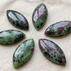 May include: Six marquise-shaped cabochons made of zoisite with ruby inclusions. Each stone features a polished, smooth surface displaying a mottled pattern of deep forest green, black, and vibrant magenta-pink spots arranged in a circular formation.