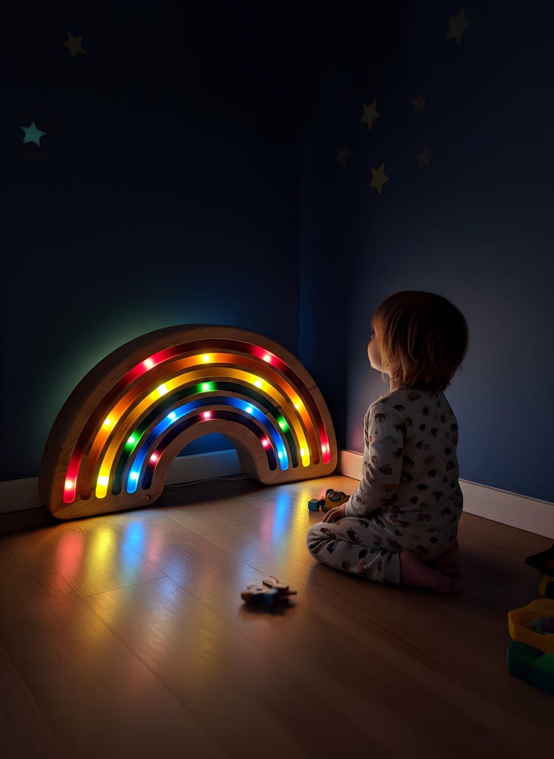 May include: Wooden rainbow-shaped night light with colorful LED lights. The rainbow is on a wooden floor in a dark room. The rainbow light is illuminating the room. A child is sitting on the floor looking at the rainbow.