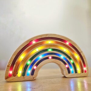 May include: Wooden rainbow-shaped night light with embedded LED lights. The rainbow features bands of red, orange, yellow, green, and blue. The lights are visible along the inner edges of each band, creating a colorful glow. The wood has a natural grain.