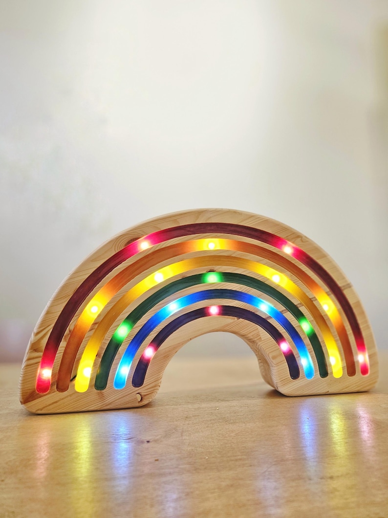 May include: Wooden rainbow-shaped night light with embedded LED lights. The rainbow features bands of red, orange, yellow, green, and blue. The lights are visible along the inner edges of each band, creating a colorful glow. The wood has a natural grain.