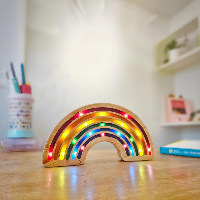 May include: Wooden rainbow-shaped night light with embedded LED lights in red, orange, yellow, green, and blue. The light is made of light-colored wood and sits on a wooden surface. The rainbow is a decorative item.