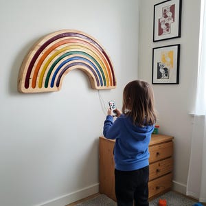 以下が含まれることがあります： 木製の虹の壁掛けアート。赤、オレンジ、黄色、緑、青の層で構成された虹のデザイン。白い壁に取り付けられています。子供がリモコンを持っています。