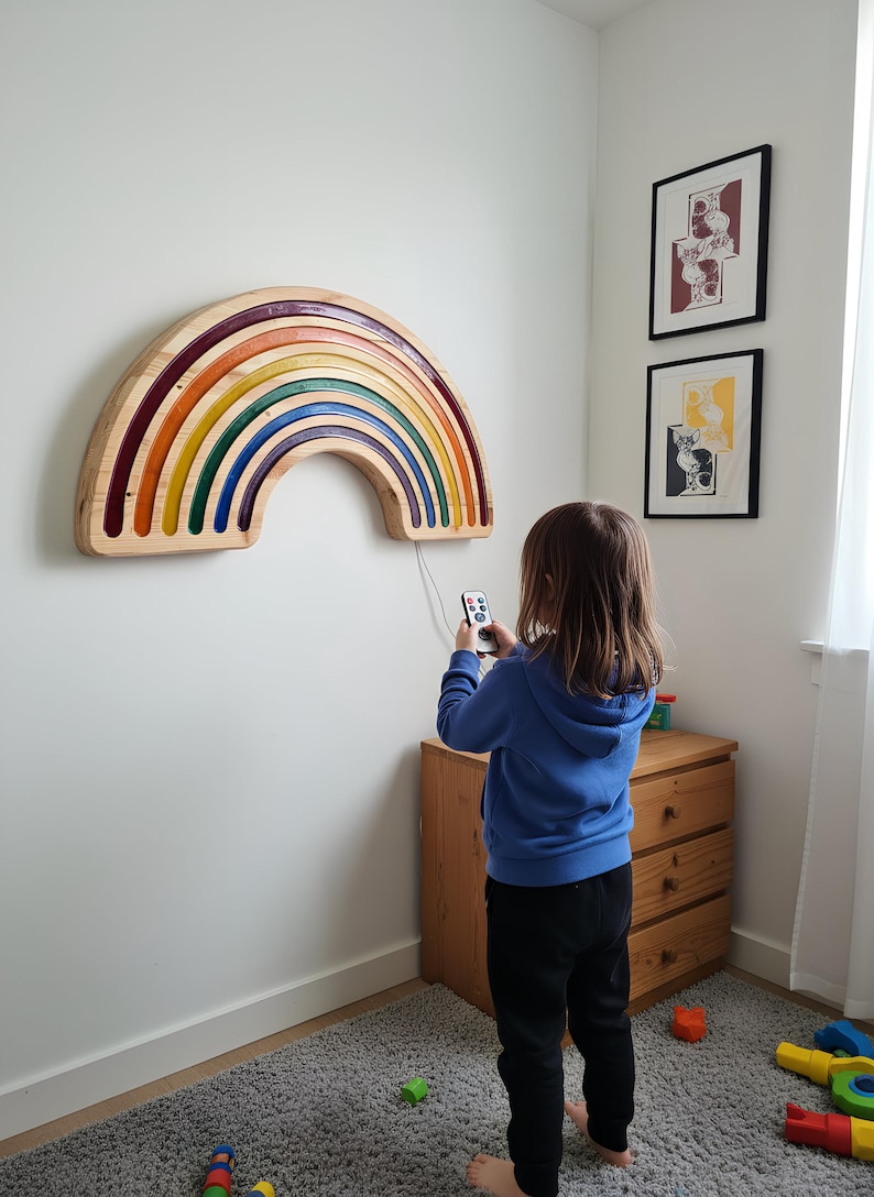 May include: Wooden rainbow wall art with a multi-colored design. The rainbow features layers of red, orange, yellow, green, and blue. The art is mounted on a white wall. A child is holding a remote control.