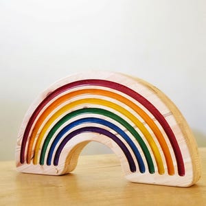 May include: Wooden rainbow toy with seven arched layers in vibrant colors: red, orange, yellow, green, blue, indigo, and violet. The toy is made of natural wood with a smooth finish, and the arches are nested together. A colorful educational toy.