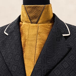 May include: A black blazer with a subtle geometric pattern and white piping on the lapels, worn over a mustard-gold high-neck garment featuring a textured, scale-like triangular panel at the collar and a smooth, lustrous finish on the chest.