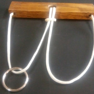 May include: A wooden puzzle with a white rope threaded through holes in the wood. The rope is tied in a knot and forms a loop. A metal ring is attached to the rope.