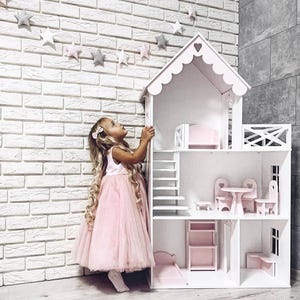 Könnte beinhalten: Ein weißes Holzpuppenhaus mit rosa Akzenten, das mehrere Zimmer, eine Treppe und Miniaturmöbel aufweist. Das Puppenhaus hat einen herzförmigen Ausschnitt auf dem Dach. Ein junges Mädchen in einem rosa Kleid steht daneben.
