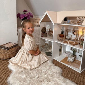 Könnte beinhalten: Ein junges Kind in einem geblümten Kleid sitzt in der Nähe eines großen, weißen Puppenhauses mit möblierten Zimmern. Das Puppenhaus hat ein Satteldach und mehrere Etagen. Die Szene spielt sich auf einem flauschigen weißen Teppich und einer gewebten Matte ab.