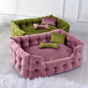 May include: Two plush dog beds. One is a dusty rose colour, and the other is olive green. Each bed has a matching cushion and a bone-shaped toy. The beds are on a light-coloured floor.