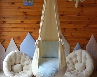 May include: A cream-colored hanging swing chair with a light blue cushion and a matching round floor cushion. The swing is suspended from the ceiling and tied with cream-colored bows. The background features a wooden wall and decorative elements.