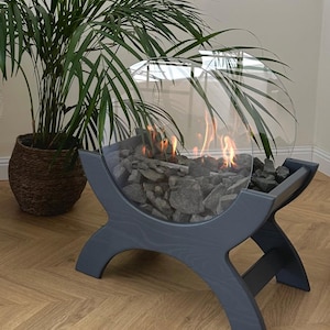May include: A modern, round, glass-enclosed fireplace with a dark grey wooden stand. The fireplace is filled with black rocks and has a warm, orange flame burning inside.