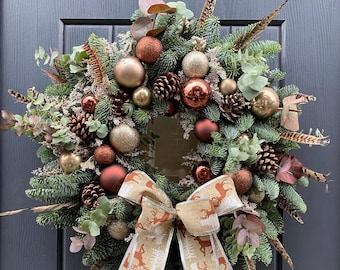 Könnte beinhalten: Ein festlicher Weihnachtskranz mit Tannenzweigen, Tannenzapfen und goldenen und kupferfarbenen Kugeln. Der Kranz ist mit einer großen Schleife mit Hirschmuster und Federn verziert.
