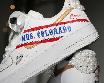 May include: White sneakers customized with pearl and rhinestone embellishments. The shoes feature red and gold accents with the text "MRS. COLORADO" and "American". The design includes a gold swoosh and white laces.