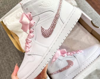May include: White high-top sneakers with a pink satin ribbon and a pink rhinestone Nike swoosh.