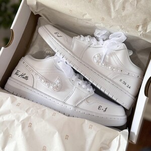 May include: A pair of white leather sneakers with pearl embellishments. The sneakers have the text "The Hulls" and "E+J" written on them, along with the date "11.09.24".