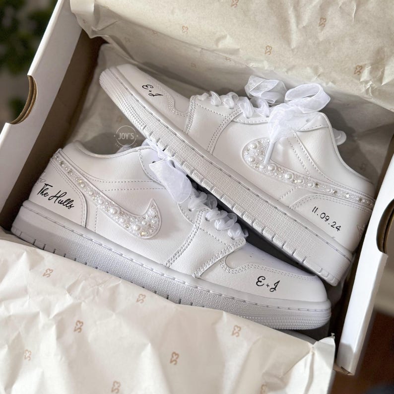 May include: A pair of white leather sneakers with pearl embellishments. The sneakers have the text "The Hulls" and "E+J" written on them, along with the date "11.09.24".
