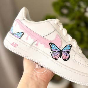 Puede incluir: Zapatillas blancas de cuero con un dise&ntilde;o de mariposa rosa y azul. Las zapatillas tienen un swoosh rosa y un dise&ntilde;o de goteo rosa. El texto "AIR" est&aacute; impreso en el lateral de la zapatilla.