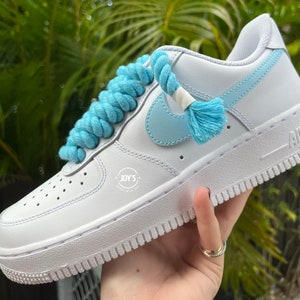 K&ouml;nnte beinhalten: Wei&szlig;e Leder-Sneaker mit niedrigem Schaft, einem hellblauen Swoosh und einer hellblauen Schn&uuml;rsenkel aus Seil. Die Schn&uuml;rsenkel sind auf einzigartige Weise mit einem hellblauen Quasten an den Enden gebunden. Die Sneaker haben eine wei&szlig;e Sohle und eine wei&szlig;e Zwischensohle. Der Text "JOY'S" ist auf der Seite des Schuhs aufgedruckt.