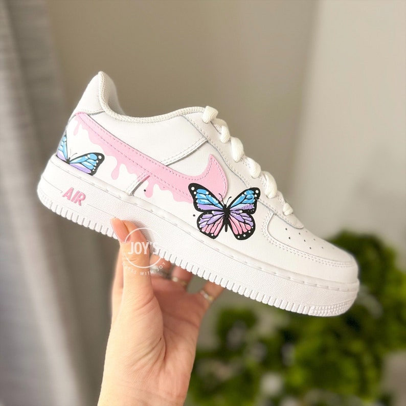 Puede incluir: Zapatillas blancas de cuero con un dise&ntilde;o de goteo rosa y dos ilustraciones de mariposas. Las mariposas est&aacute;n pintadas en tonos de azul, rosa y morado. Las zapatillas dicen "AIR" en el lateral.