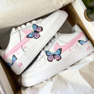 Puede incluir: Un par de zapatillas blancas con dise&ntilde;os de mariposas rosas y azules. Las zapatillas tienen un swoosh rosa con pintura rosa goteando y la palabra "AIR" en el lateral.