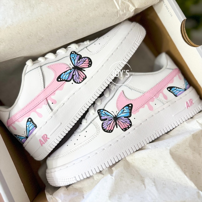 Puede incluir: Un par de zapatillas blancas con dise&ntilde;os de mariposas rosas y azules. Las zapatillas tienen un swoosh rosa con pintura rosa goteando y la palabra "AIR" en el lateral.
