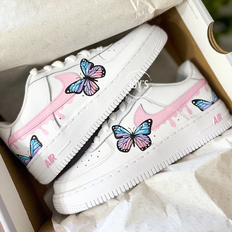 Puede incluir: Un par de zapatillas blancas con dise&ntilde;os de mariposas rosas y azules. Las zapatillas tienen un swoosh rosa y la palabra "AIR" en el lateral.