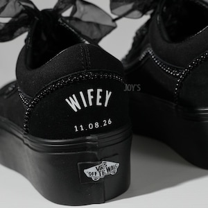 May include: Black platform sneakers with a decorative beaded trim and sheer black bows. The word "WIFEY" and the date "11.08.26" are printed in white on the heel. The Vans logo is visible on the side.