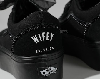 May include: Black platform sneakers with a decorative beaded trim and sheer black bows. The word "WIFEY" and the date "11.08.26" are printed in white on the heel. The Vans logo is visible on the side.