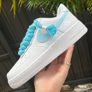 K&ouml;nnte beinhalten: Wei&szlig;e Leder-Sneaker mit niedrigem Schaft, einem hellblauen Swoosh und Schn&uuml;rsenkeln. Die Schn&uuml;rsenkel sind mit einem hellblauen Seil gebunden und haben einen hellblauen Quasten. Der Text "JOY'S" ist auf der Seite des Schuhs aufgedruckt.