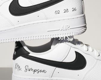 May include: A pair of white sneakers with black Nike swoosh logos and black heel tabs. The top shoe features the date 02.28.26 in black script. The bottom shoe features the text Mr. Simpson in black script. Both shoes have white soles with AIR text.