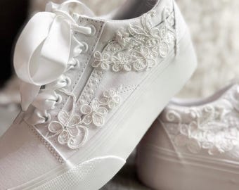 May include: White canvas sneakers with floral lace appliqués and satin ribbon laces. The platform soles are white, and the shoes have a classic lace-up design. The floral details add a touch of elegance to the casual style.
