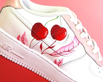 May include: White leather sneaker with a cherry design. The shoe features a pink heel tab and a painted design of two red cherries with green stems and pink leaves. The word "AIR" is visible on the side of the sole.