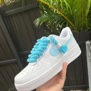 K&ouml;nnte beinhalten: Wei&szlig;e Leder-Sneaker mit niedrigem Schaft, einem hellblauen Swoosh und Schn&uuml;rsenkeln. Die Schn&uuml;rsenkel bestehen aus einem dicken, blauen Seil. Die Sneaker haben eine wei&szlig;e Sohle und ein kleines wei&szlig;es Etikett an der Seite mit der Aufschrift "JOY'S".