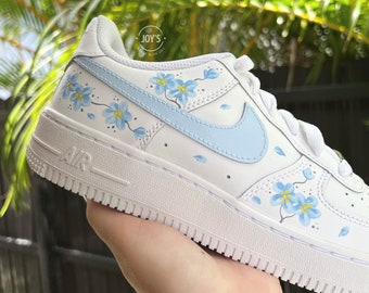 May include: White leather sneakers with a light blue swoosh and blue flower design. The sneakers have the word "AIR" printed on the side.