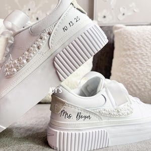 May include: White platform sneakers with pearl and rhinestone embellishments. The sneakers have a ribbon detail and the text "10.13.25" and "Mrs. Boyer". These custom shoes are perfect for a wedding or special occasion.