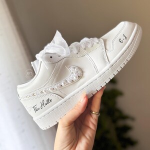 May include: White leather low-top sneakers with a pearl-embellished swoosh and the text "The Hulls" and "3-4" written on the side.