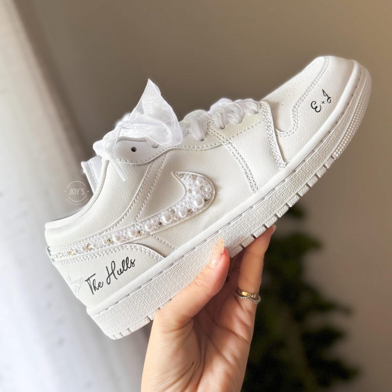 May include: White leather low-top sneakers with a pearl-embellished swoosh and the text "The Hulls" and "3-4" written on the side.